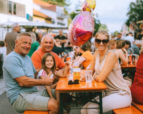 Auf dem Bild sieht man eine Familie beim Traunreuter Stadtfest an einem Biertisch in der Kantstraße sitzen: zwei Männer, ein Kind mit einem pinken Luftballon und eine Frau.